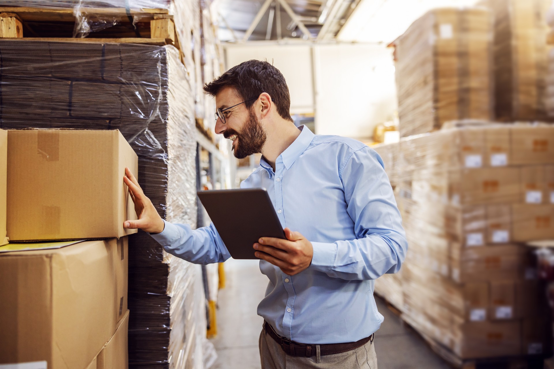 Young smiling supervisor standing in warehouse with tablet in hands and checking out order for customers.