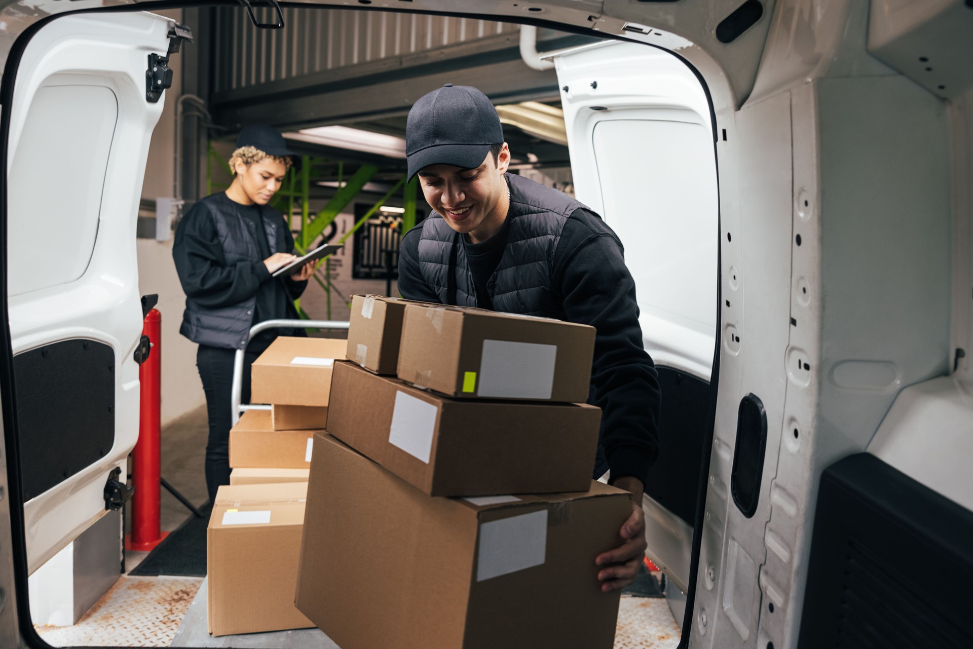 Smiling male courier loads cardboard boxes from a cart into the car while coworker checking information on digital tablet in warehouse